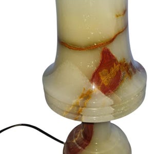May include: A decorative table lamp made of translucent stone. The lamp features a rounded base, a flared midsection, and a cylindrical top. The stone has a creamy white color with reddish-brown and gold veining. A black power cord extends from the base.