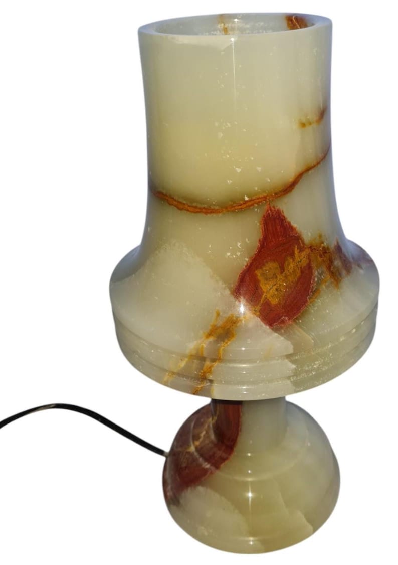 May include: A decorative table lamp made of translucent stone. The lamp features a rounded base, a flared midsection, and a cylindrical top. The stone has a creamy white color with reddish-brown and gold veining. A black power cord extends from the base.