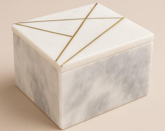 Op de afbeelding: Een rechthoekige marmeren doos met een deksel. De doos is wit met grijze adering en het deksel heeft een geometrisch goudkleurig ontwerp. De doos is een decoratief opbergitem.
