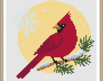May include: Cross-stitch artwork of a vibrant red cardinal bird perched on a snow-covered branch with green pine needles. The bird is set against a yellow circle with a snowflake and small white dots, all within a framed square.