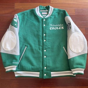 May include: Green and gray Philadelphia Eagles letterman jacket. The jacket features the team name and logo on the front, with white elbow patches and striped trim. The NFL logo is on the sleeve.