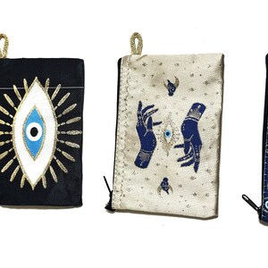 May include: Four small, rectangular pouches with gold-colored accents and zippers. The pouches feature embroidered designs of the evil eye, stars, and hands. Colors include blue, black, and cream.