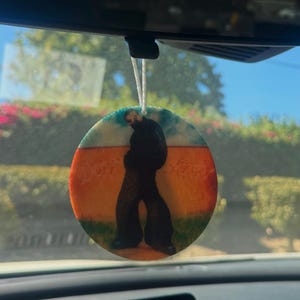 May include: A circular car air freshener featuring a silhouette of a person in black clothing against a backdrop of orange and blue. The air freshener is hanging from a white string, visible through a car's rearview mirror.