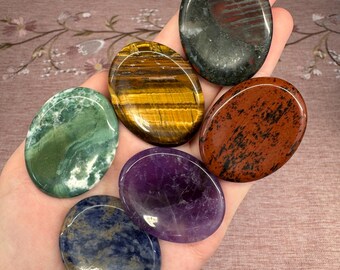 May include: A collection of seven polished, oval-shaped gemstones in various colors. The stones include green, gold, purple, blue, red, and brown hues. The gemstones are smooth and appear to be held in a hand.