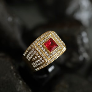 Può includere: Un anello dorato con una gemma rossa quadrata al centro. L'anello è incastonato con piccole gemme trasparenti attorno alla pietra centrale e lungo la fascia, creando un effetto scintillante. L'anello è posto su uno sfondo scuro.