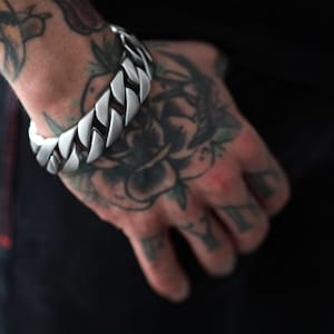 Può includere: Un bracciale a catena a maglia larga color argento. Il bracciale è composto da maglie arrotondate e interconnesse. Il bracciale è indossato su un polso tatuato. È un gioiello audace.