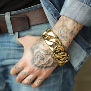 May include: A gold-colored, chunky chain bracelet. The bracelet is made of interlocking links and is worn on a wrist with tattoos. The person is wearing a denim jacket and jeans.