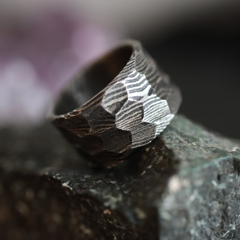 Peut inclure: Gros plan sur une bague gris fonc&eacute; textur&eacute;e avec un motif g&eacute;om&eacute;trique. La bague pr&eacute;sente une surface &agrave; facettes avec un motif de lignes claires et sombres. La bague repose sur une surface sombre et textur&eacute;e.