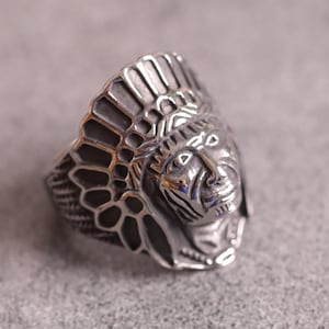 Puede incluir: Un anillo plateado con una detallada representación del rostro y el tocado de un jefe nativo americano. El anillo tiene intrincadas tallas y un diseño audaz. El anillo probablemente está hecho de una aleación de metal.