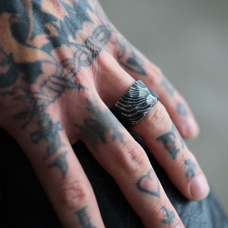 Peut inclure: Gros plan d'une bague argent&eacute;e avec un motif textur&eacute; en forme de vagues. La bague est de forme carr&eacute;e et port&eacute;e au doigt. La main est tatou&eacute;e.