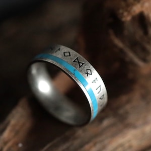 Może przedstawiać: Srebrny pierścionek z turkusową wstawką i wygrawerowanymi symbolami runicznymi. Pierścionek ma zużyty, antyczny wygląd. Opaska ma jasnoniebieski pasek. To biżuteria.