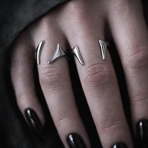 May include: A close-up of a hand wearing three silver rings. The rings have a claw-like design, with sharp, pointed elements. The nails are painted black, creating a contrast with the silver rings.