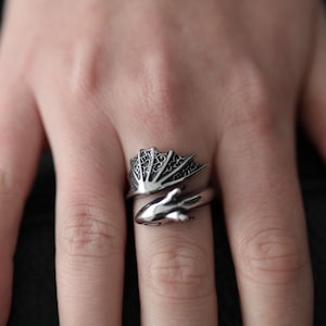 May include: A silver-colored ring with a dragon wing design. The ring wraps around the finger, featuring a detailed wing and a dragon head. The ring is made of a shiny metal and is shown on a person's finger.