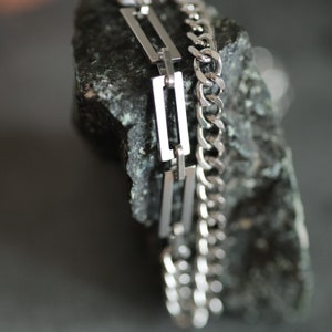 Puede incluir: Primer plano de dos collares plateados. Uno presenta un diseño de cadena de eslabones, el otro eslabones rectangulares. Los collares se exhiben sobre una roca oscura y texturizada, contrastando el metal brillante con la superficie rugosa.