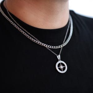 May include: Silver-toned necklace set featuring a curb chain and a pendant with a compass rose design. The pendant has a star in the center and Roman numerals around the edge. The necklace is displayed against a black shirt.