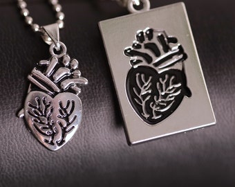 Può includere: Due collane con ciondoli a forma di cuore argentati. Un ciondolo è a forma di cuore, l'altro rettangolare. Entrambi presentano un design dettagliato del cuore anatomico. Le collane sono appese a catene a sfera, su sfondo scuro.