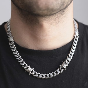 May include: A silver chain necklace with a curb link design. The necklace has barbed wire accents at intervals along the chain. The necklace is worn against a black top.