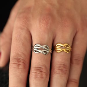 Op de afbeelding: Twee ringen, één zilverkleurig en één goudkleurig, elk met een knoopontwerp. De ringen worden op een hand getoond, waarbij hun ingewikkelde details en metallic glans worden benadrukt. Het knoopontwerp voegt een unieke touch toe aan de sieraden.