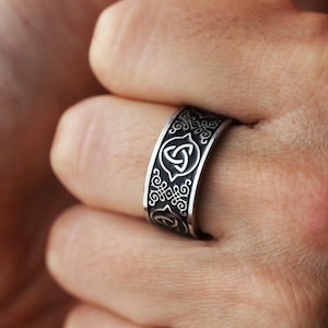 Könnte beinhalten: Ein silberfarbener Ring mit einem keltischen Knotenmuster. Der Ring hat aufwendige Muster und ein breites Band. Der zentrale Knoten ist von einem dekorativen Rand umgeben. Der Ring wird am Finger getragen.