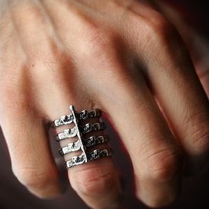 Puede incluir: Un anillo plateado con un diseño único que presenta múltiples bandas conectadas por un elemento vertical central. El anillo tiene un acabado texturizado, posiblemente oxidado, y se lleva en un dedo.