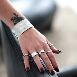 Könnte beinhalten: Eine Sammlung von Silberringen in verschiedenen Designs, darunter ein V-förmiger Ring, und ein breites silbernes Armband. Der Schmuck wird auf einer Hand mit dunklem Nagellack und einem floralen Tattoo am Handgelenk präsentiert.