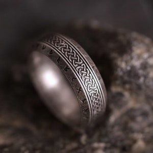 Może przedstawiać: Srebrny pierścień z celtyckim wzorem węzła i wygrawerowanymi runami. Pierścień ma szeroką obręcz ze szczegółowym wzorem pośrodku. Tło jest rozmyte, ciemnoszare.
