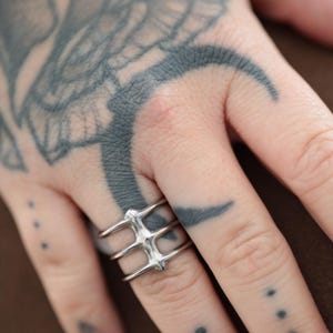 May include: A silver ring with a unique, geometric design. The ring features three parallel bands connected by vertical bars, creating a cage-like structure. The ring is worn on a hand with intricate tattoos.