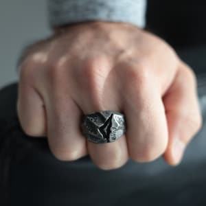 May include: A textured, dark gray ring with a jagged, cracked design. The ring is worn on a finger, showcasing its unique, rugged aesthetic. The ring's surface has a rough, stone-like appearance, adding to its distinctive character.