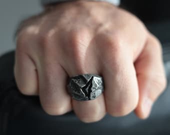 Op de afbeelding: Een donkergrijze ring met een getextureerd, gebarsten ontwerp. De ring wordt aan een vinger gedragen en benadrukt zijn unieke, robuuste esthetiek. Het oppervlak van de ring heeft een ruwe, steenachtige uitstraling.