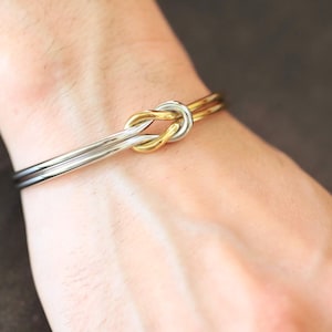 Könnte beinhalten: Ein zweifarbiges Armband mit einem Knotendesign. Das Armband besteht aus silber- und goldfarbenem Metall. Der Knoten ist der Mittelpunkt, wobei sich das silberne Band um das Handgelenk krümmt.