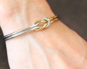 以下が含まれることがあります： ノットデザインが特徴のツートンカラーのブレスレット。ブレスレットはシルバーとゴールドカラーの金属製です。ノットが焦点で、シルバーのバンドが手首に沿ってカーブしています。