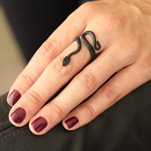 Puede incluir: Un anillo negro con forma de serpiente se enrolla alrededor de un dedo. El anillo tiene un diseño detallado de serpiente con un cuerpo texturizado y una pequeña cabeza. La mano tiene esmalte de uñas burdeos.