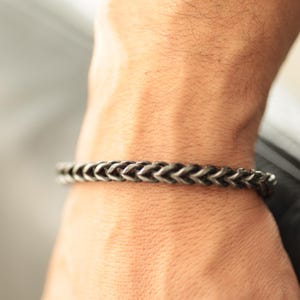 Op de afbeelding: Een zilverkleurige armband met een gevlochten kettingpatroon. De metalen armband heeft een getextureerde afwerking. De armband is ongeveer 20 cm lang en wordt om de pols gedragen.