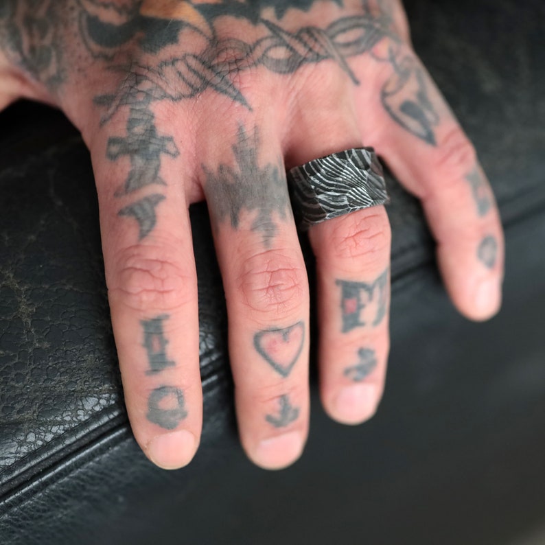Peut inclure: Gros plan d'une bague argent&eacute;e avec un motif textur&eacute;. La bague est port&eacute;e sur un doigt tatou&eacute; de divers motifs, dont un c&oelig;ur, une ancre et d'autres symboles. La surface de la bague refl&egrave;te la lumi&egrave;re, soulignant ses d&eacute;tails.