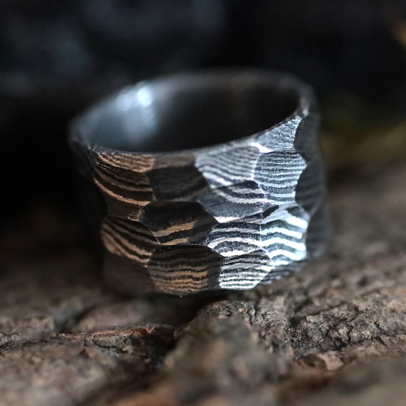 Peut inclure: Gros plan sur une bague textur&eacute;e gris fonc&eacute;. La bague pr&eacute;sente un motif unique de lignes et de formes g&eacute;om&eacute;triques. La bague semble &ecirc;tre en m&eacute;tal et a un aspect robuste et artisanal. La bague est un bijou.