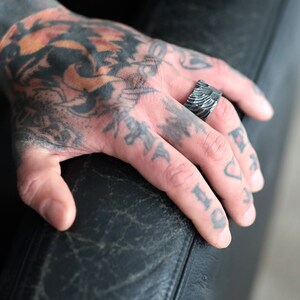 Peut inclure: Une bague argent&eacute;e textur&eacute;e avec un motif unique. La bague est port&eacute;e sur un doigt tatou&eacute;. Le design de la bague pr&eacute;sente des lignes parall&egrave;les, cr&eacute;ant une texture visuelle distinctive. La bague est un bijou.