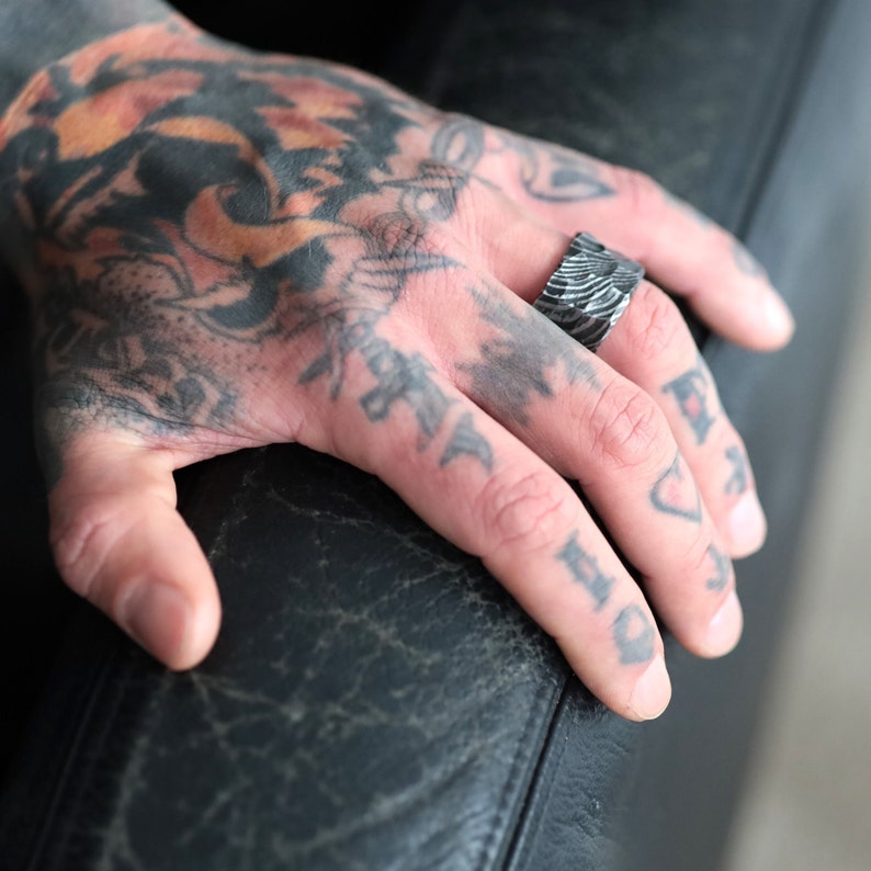 Peut inclure: Une bague argent&eacute;e textur&eacute;e avec un motif unique. La bague est port&eacute;e sur un doigt tatou&eacute;. Le design de la bague pr&eacute;sente des lignes parall&egrave;les, cr&eacute;ant une texture visuelle distinctive. La bague est un bijou.
