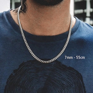 Peut inclure: Un collier chaîne gourmette argenté, de 7 mm de large et 55 cm de long. Le collier est porté par une personne portant un t-shirt bleu foncé. Le texte "7mm - 55cm" est visible.
