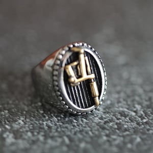 Op de afbeelding: Zilverkleurige ring met een goudkleurig tattoo-machineontwerp in een zwart ovaal. De ring heeft een gepolijste afwerking en een getextureerde rand. De tattoo-machine is een prominent kenmerk.
