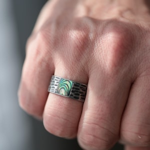 May include: A silver ring with a textured band and a square abalone inlay. The abalone inlay features iridescent green, blue, and white colors. The ring is shown on a hand.