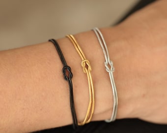 Puede incluir: Tres pulseras finas en negro, dorado y plateado, cada una con un diseño de nudo. Las pulseras son delgadas y elegantes, ideales para combinar o usar individualmente. La imagen muestra las pulseras en una muñeca, resaltando su estilo elegante y minimalista.