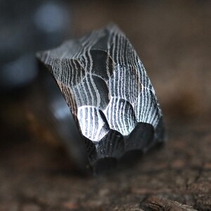 Peut inclure: Gros plan sur une bague en m&eacute;tal sombre textur&eacute;. La bague pr&eacute;sente un motif unique de lignes ondul&eacute;es et un design g&eacute;om&eacute;trique &agrave; facettes. La surface de la bague refl&egrave;te la lumi&egrave;re, soulignant les d&eacute;tails complexes du travail du m&eacute;tal. La bague est un bijou.