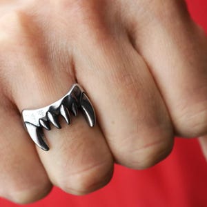 Puede incluir: Un anillo plateado con forma de colmillos de vampiro. El anillo presenta cinco dientes puntiagudos, con un diseño ligeramente curvado. El anillo se lleva en un dedo, con un fondo rojo.