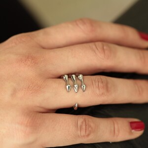 Puede incluir: Un anillo plateado con un diseño único. Presenta tres elementos en forma de lágrima que apuntan hacia arriba en el dedo y un solo elemento en forma de lágrima en el lateral. El anillo se muestra en una mano con esmalte de uñas rojo.