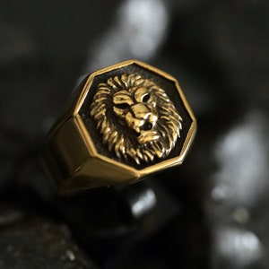 May include: A gold-colored ring with a detailed lion's head design. The ring has an octagonal face with a raised lion's head in the center. The lion's mane and facial features are intricately carved. The ring is set against a dark background.
