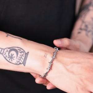May include: A silver-coloured bracelet featuring a barbed wire design. The bracelet is worn on a wrist with a tattoo of a decorative design and the number 22. The bracelet is made of metal and has sharp, pointed barbs.