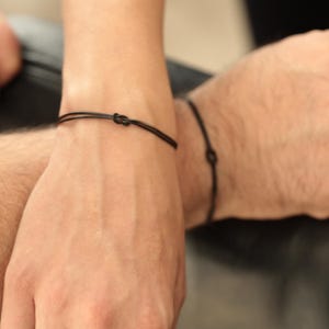 May include: Black cord bracelets with a knot design. The bracelets are simple and minimalist, suitable for everyday wear. The bracelets are shown on two wrists.