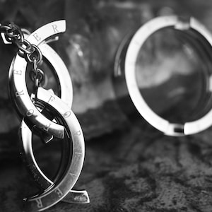 May include: A silver-toned keychain with interlocking curved segments, each inscribed with the word "FOREVER". The keychain is connected to a chain and sits next to a silver ring. The background is a textured, dark surface.