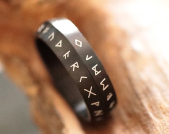 May include: A black ring with etched white runic symbols. The ring is made of a dark, possibly metal material, and the runes are evenly spaced around the band. The ring is displayed on a piece of light brown wood.