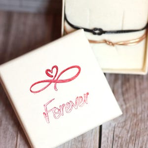 以下が含まれることがあります： 蓋に赤いハート付きの無限大シンボルと「Forever」の文字がプリントされた白い正方形のジュエリーボックス。開いた箱の中には、黒とローズゴールド調の細いコードブレスレットが2本入っています。